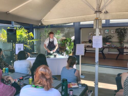 A chef wearing a white shirt and striped blue apron is standing in the corner of a sunny courtyard with a hot plate and cooking ingredients on a table in front of him. A crowd of people are watching him cook