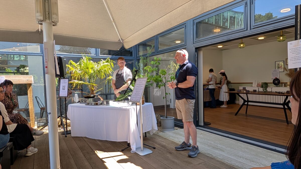 Two men stand in a sunny courtyard, one is wearing shorts and a T-shirt and talking to the crowd who are sitting in front of him. Next to him is the chef in a white shirt and blue striped apron